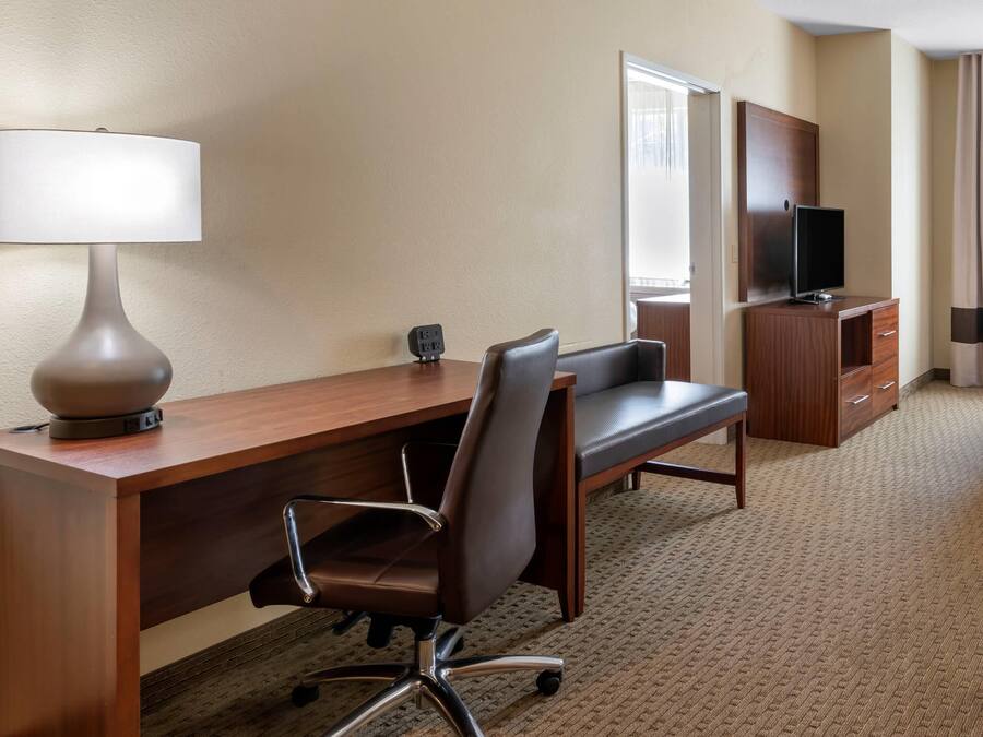 A hotel room with a wooden desk, a chair, a lamp, a television, a dresser, and a bed.