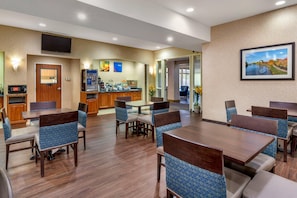 Un comedor de hotel con mesas de madera y sillas con cojines azules, una kitchenette y un televisor montado en la pared.