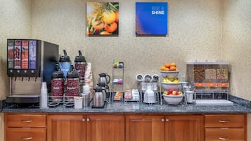 A hotel breakfast area with a coffee machine, coffee dispensers, fruit bowls, and a juice station.
