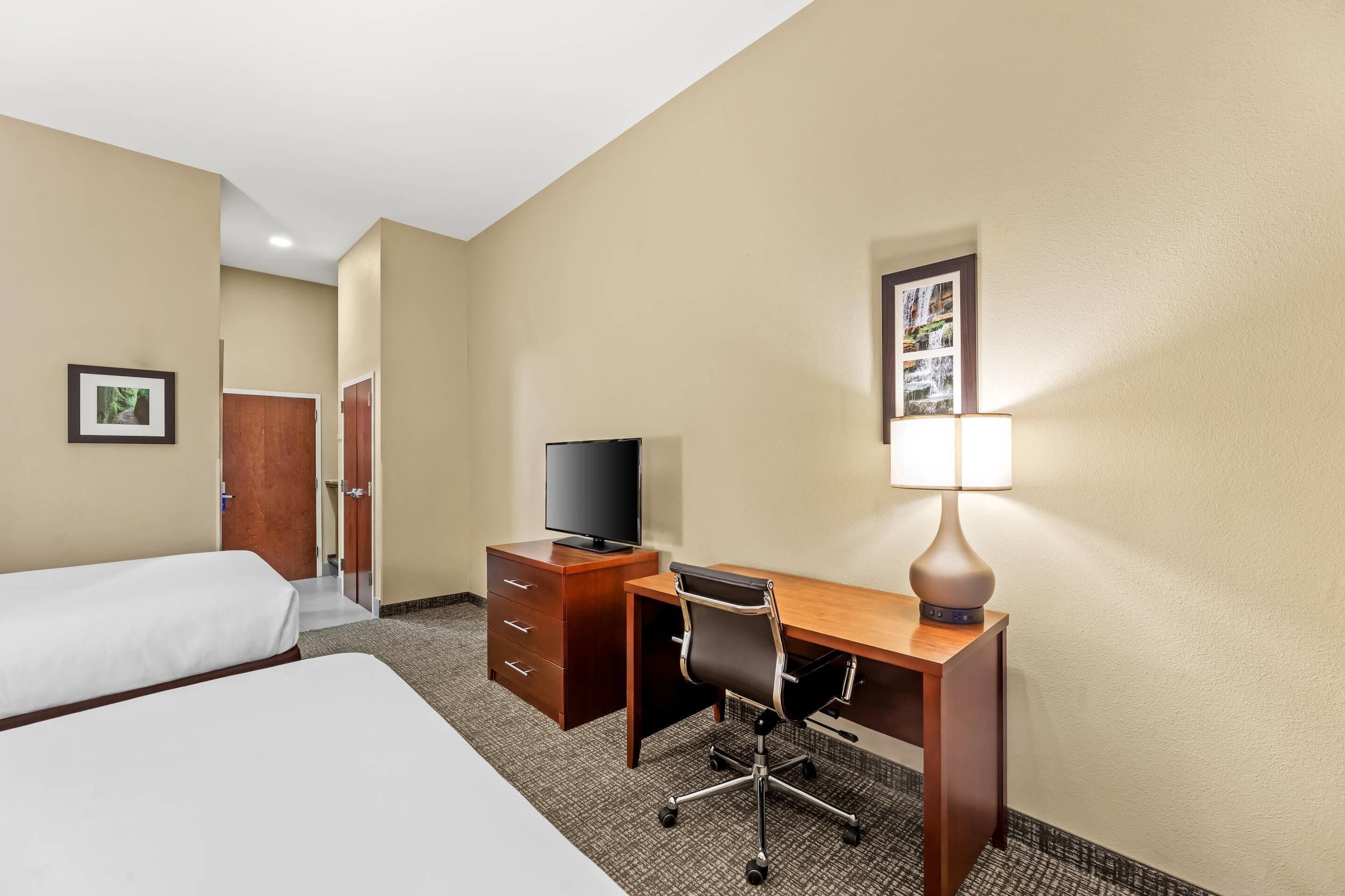 Habitación de hotel con dos camas, un escritorio con silla, un televisor, una lámpara y un cuadro en la pared.