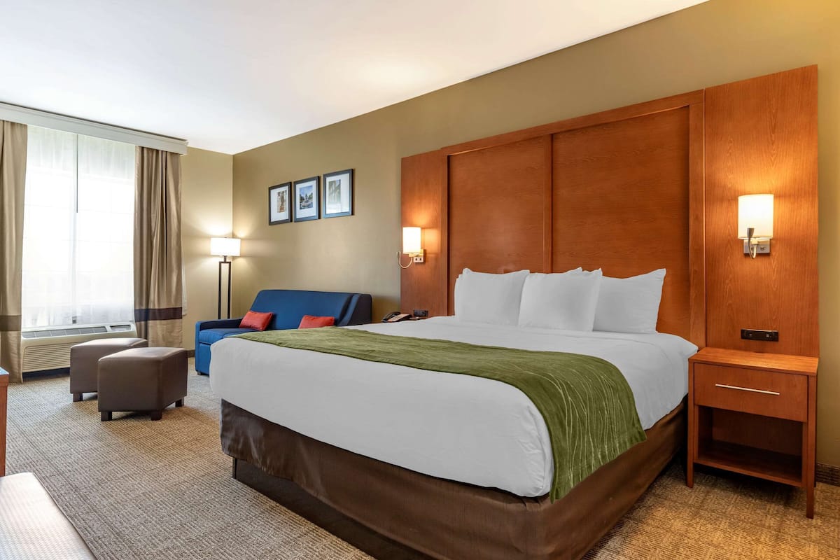 A hotel room with a large bed, a wooden headboard, two bedside tables, a desk, a chair, and a window with curtains.