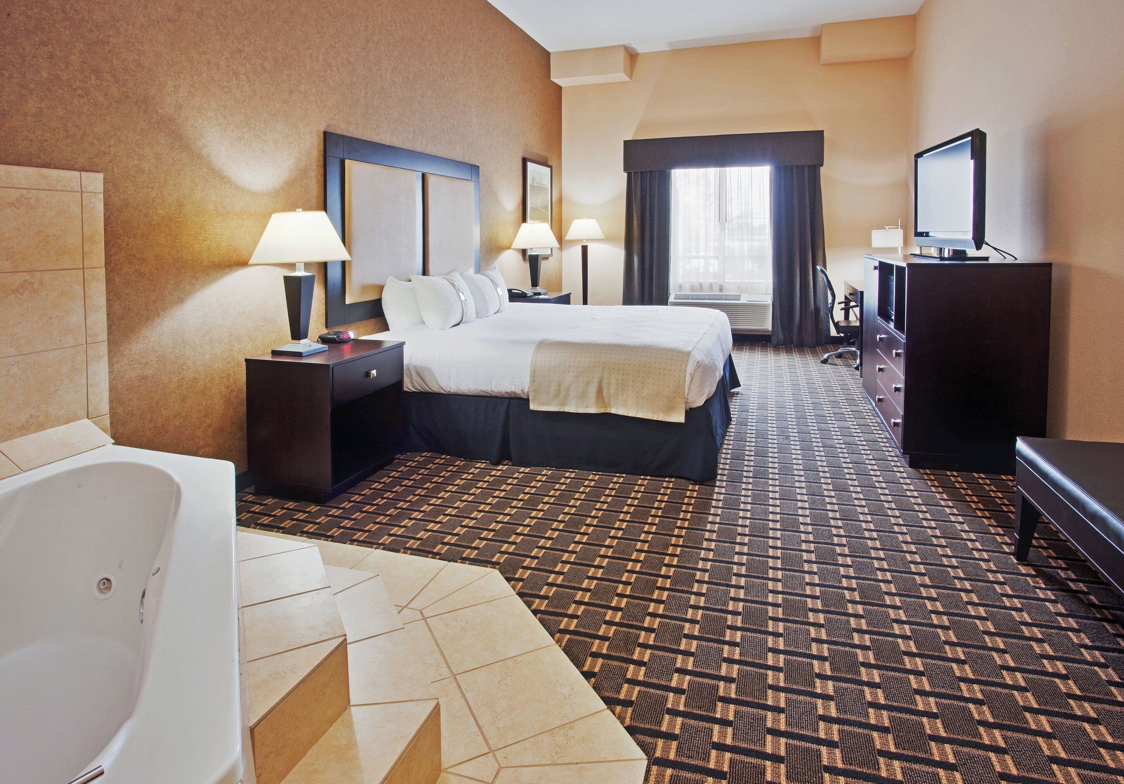 A hotel room with a bed, bedside tables, a desk, a chair, a TV, a dresser, and a bathroom with a bathtub and toilet.