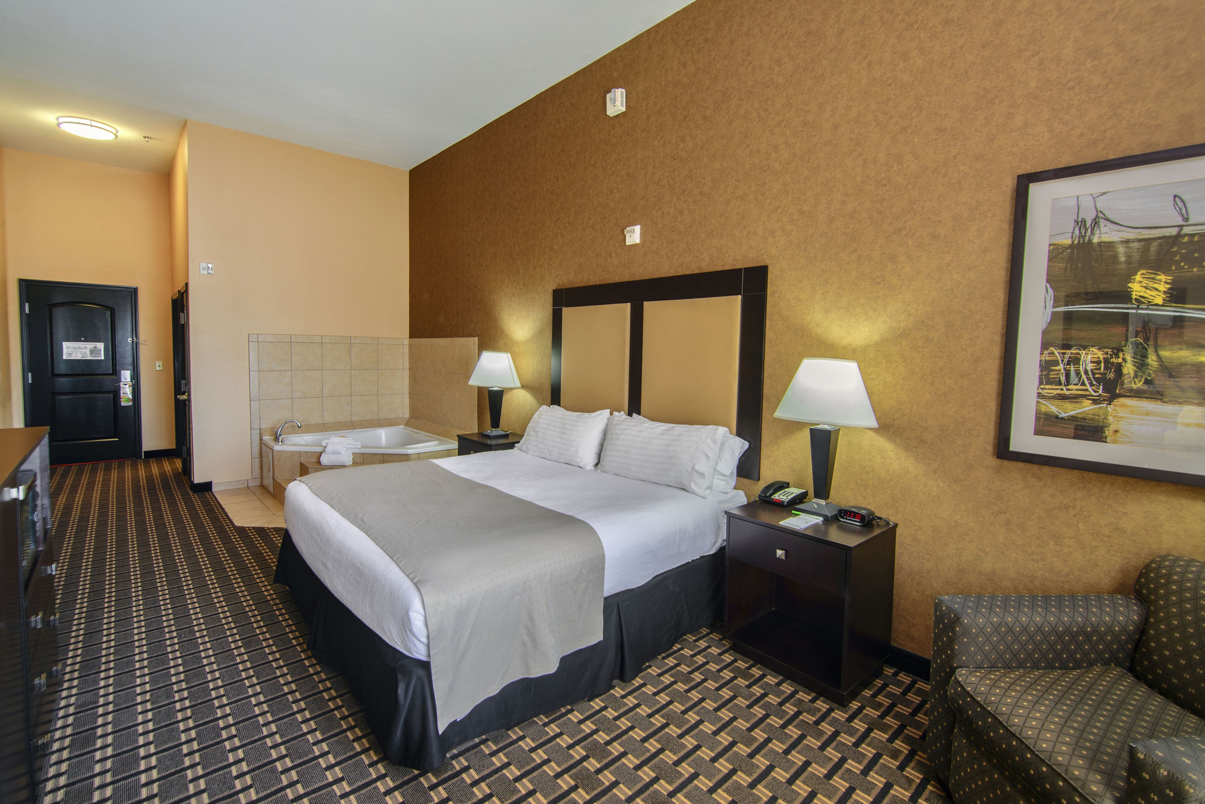 A hotel room with a bed, two bedside tables with lamps, a small desk with a telephone, a chair, and a bathroom with a sink and mirror.