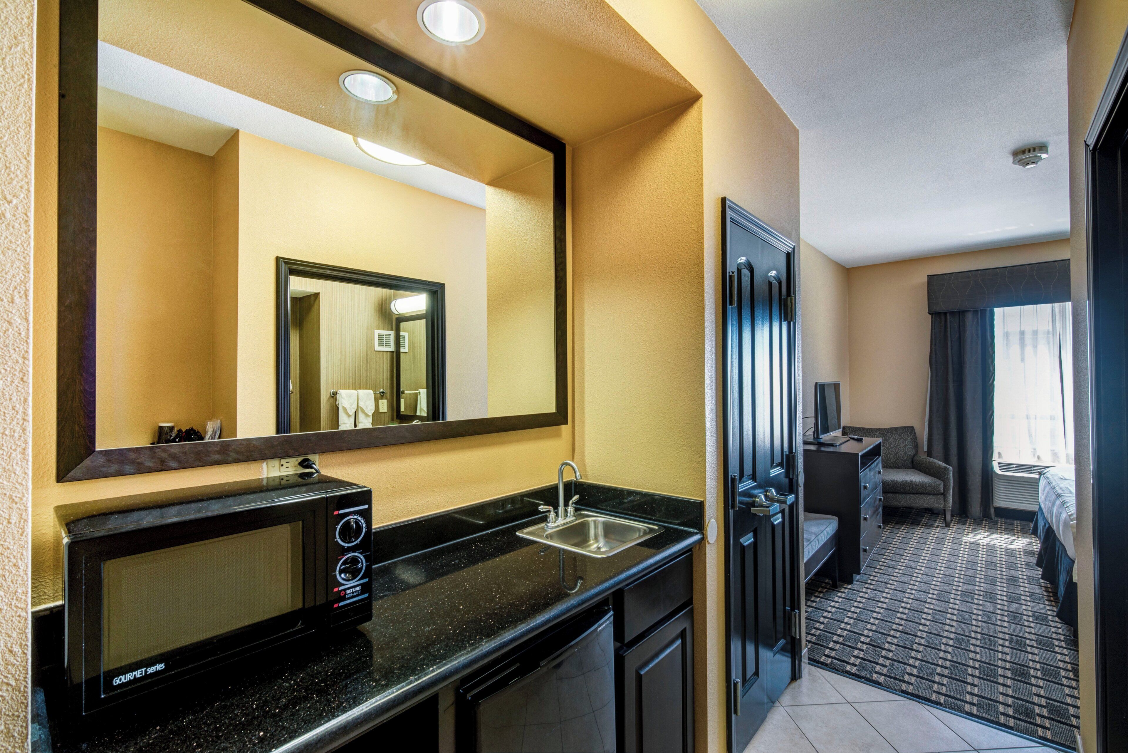 Habitación de hotel con kitchenette, un espejo grande, un lavamanos, un microondas y una puerta que da a un dormitorio.