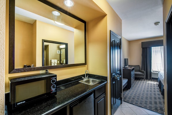 A hotel room with a kitchenette, a large mirror, a sink, a microwave, and a door leading to a bedroom.