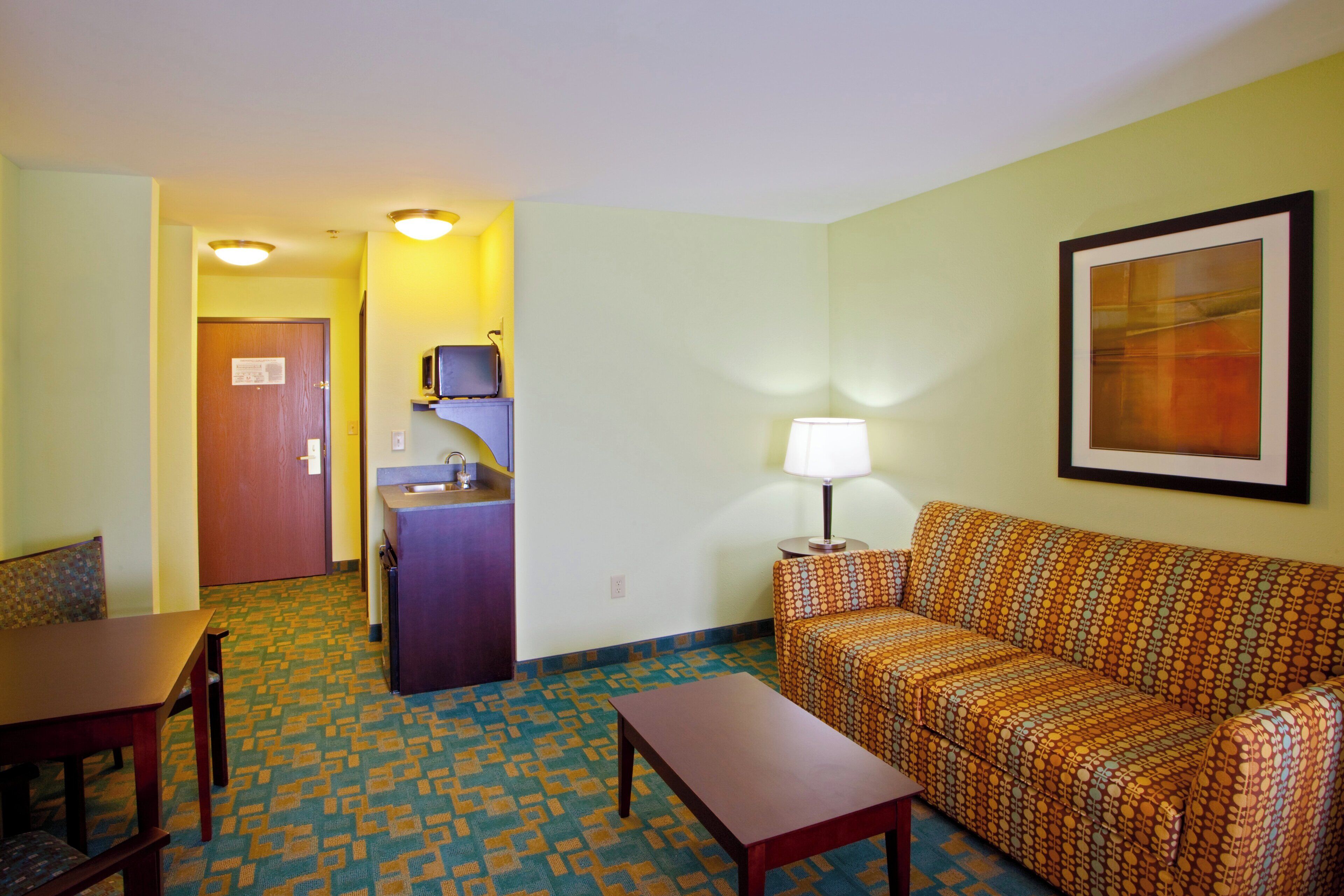 Una habitación de hotel con sofá, una mesita, escritorio, kitchenette y una puerta que da a otra habitación.