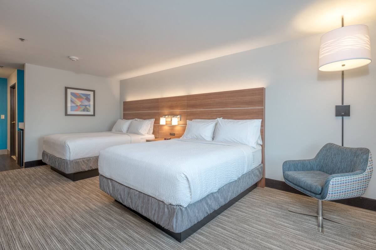 A hotel room with two beds, a blue armchair, a lamp, and a framed picture on the wall.