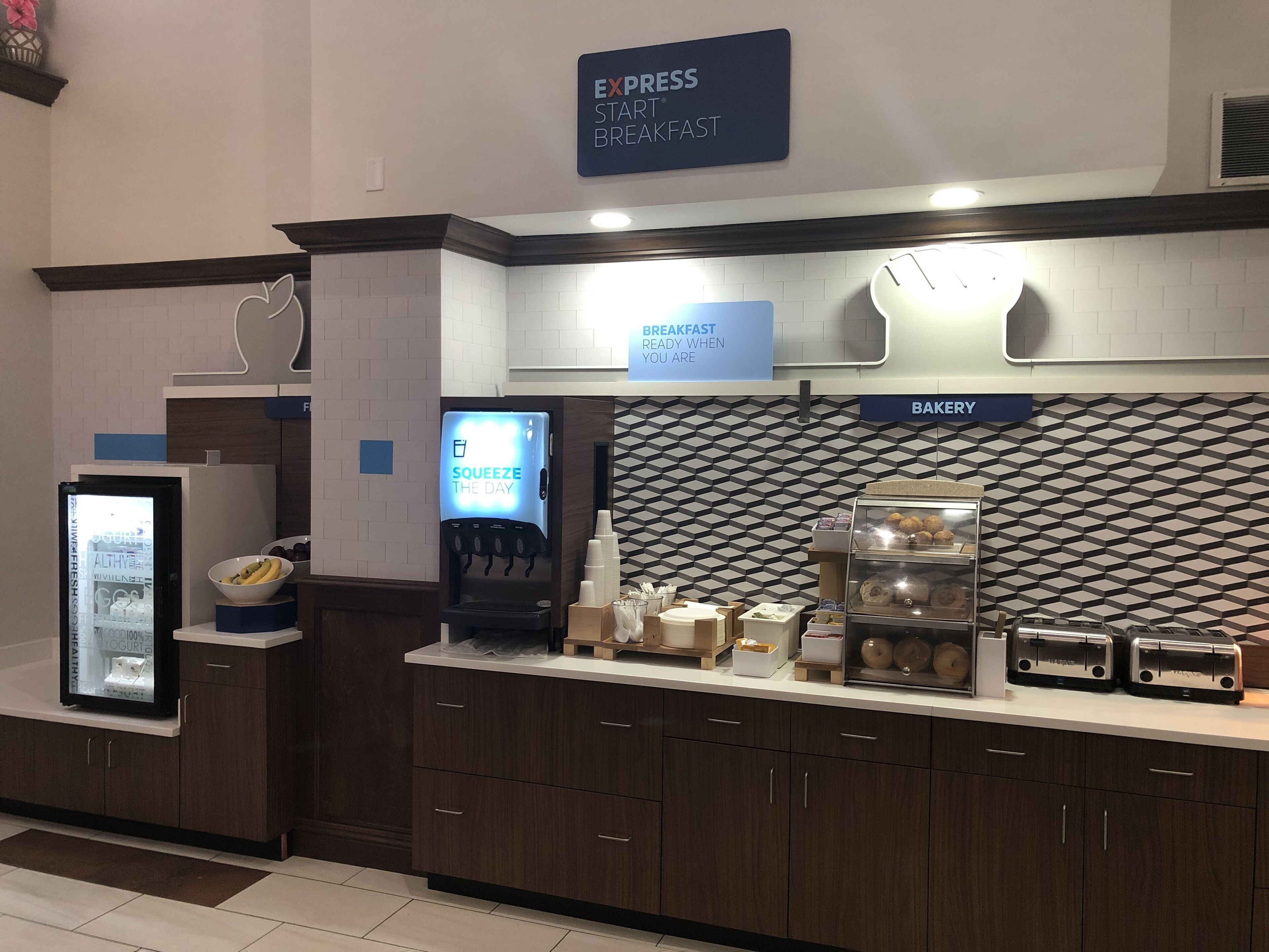 A breakfast area with a sign for 'Express Start Breakfast', a beverage dispenser, a bakery section, and a refrigerator with a yogurt display.