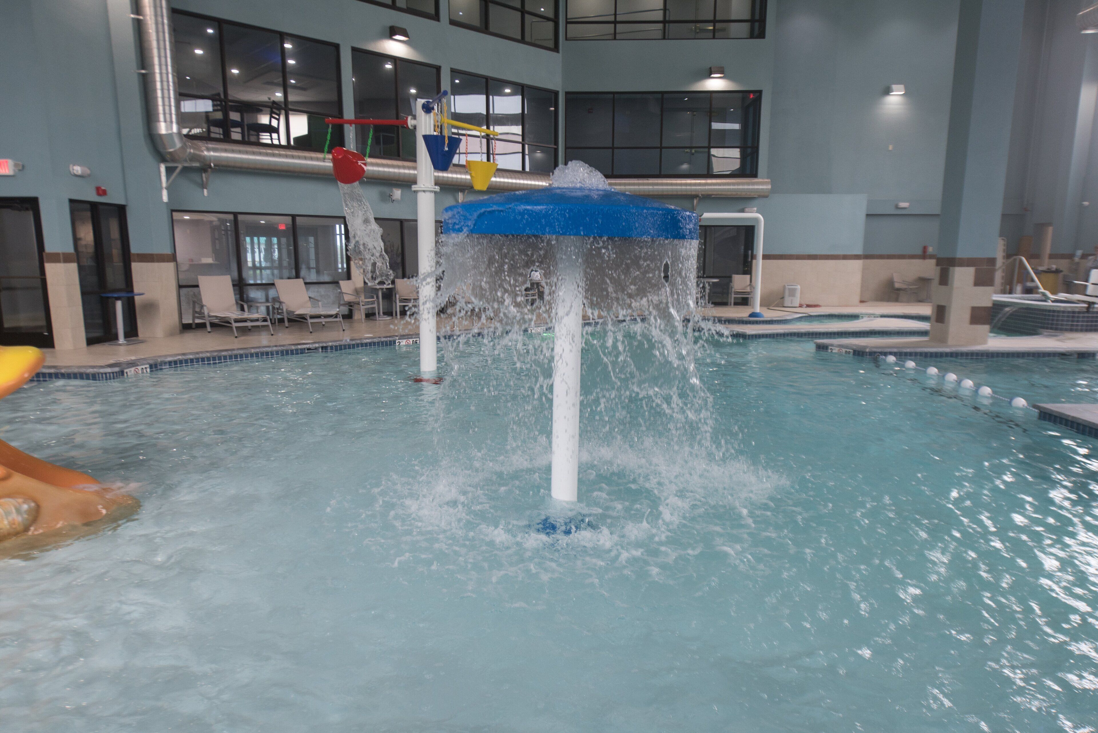 An indoor swimming pool with a water fountain feature, multiple lanes, and a seating area with chairs.