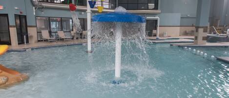An indoor swimming pool with a water fountain feature, multiple lanes, and a seating area with chairs.