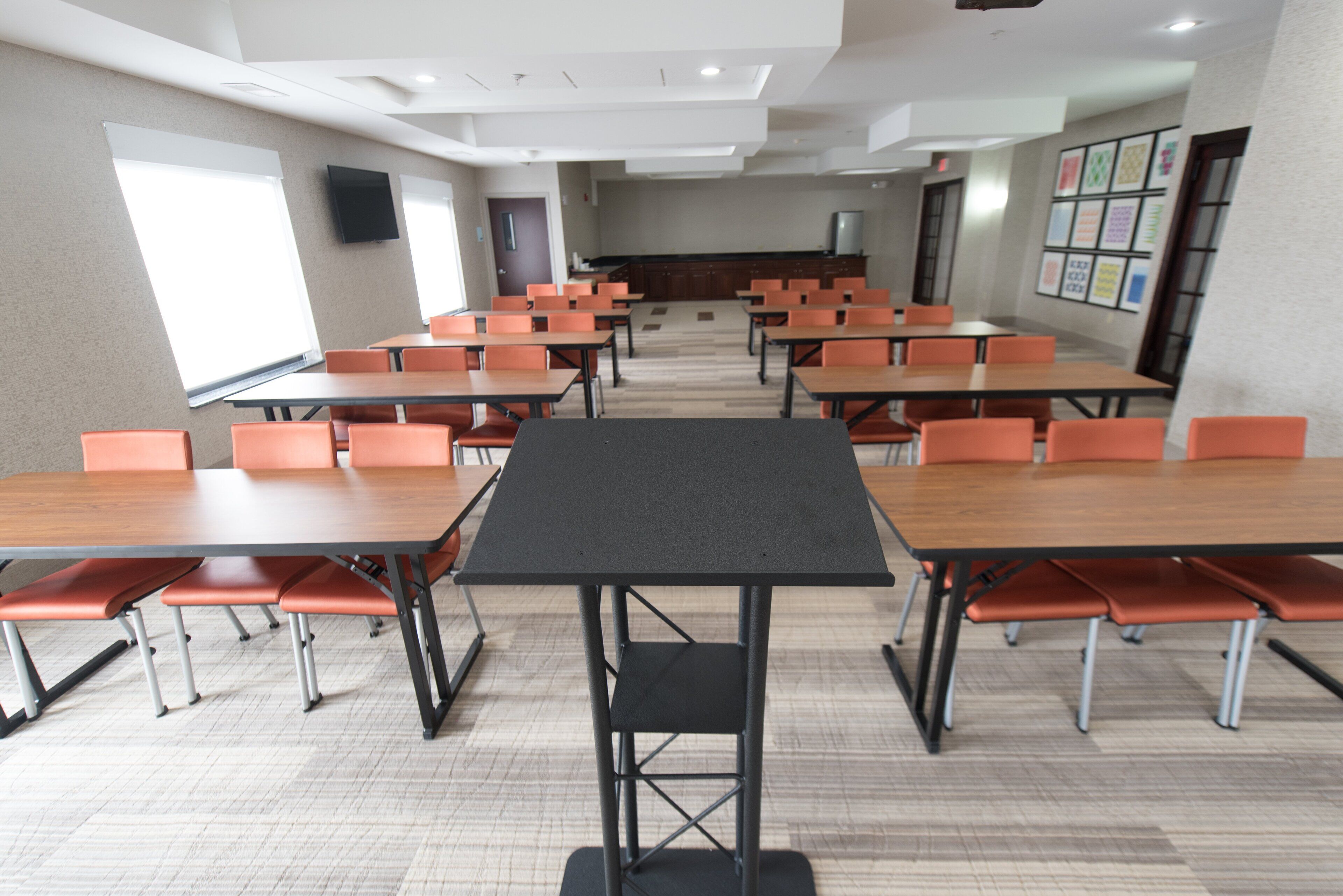 A conference room with multiple tables and chairs, a projector screen, and a kitchen area in the background.