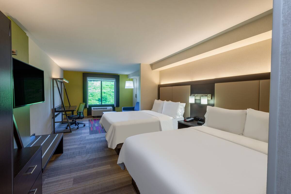 A hotel room with two beds, a desk, a TV, and a window with a view of greenery.