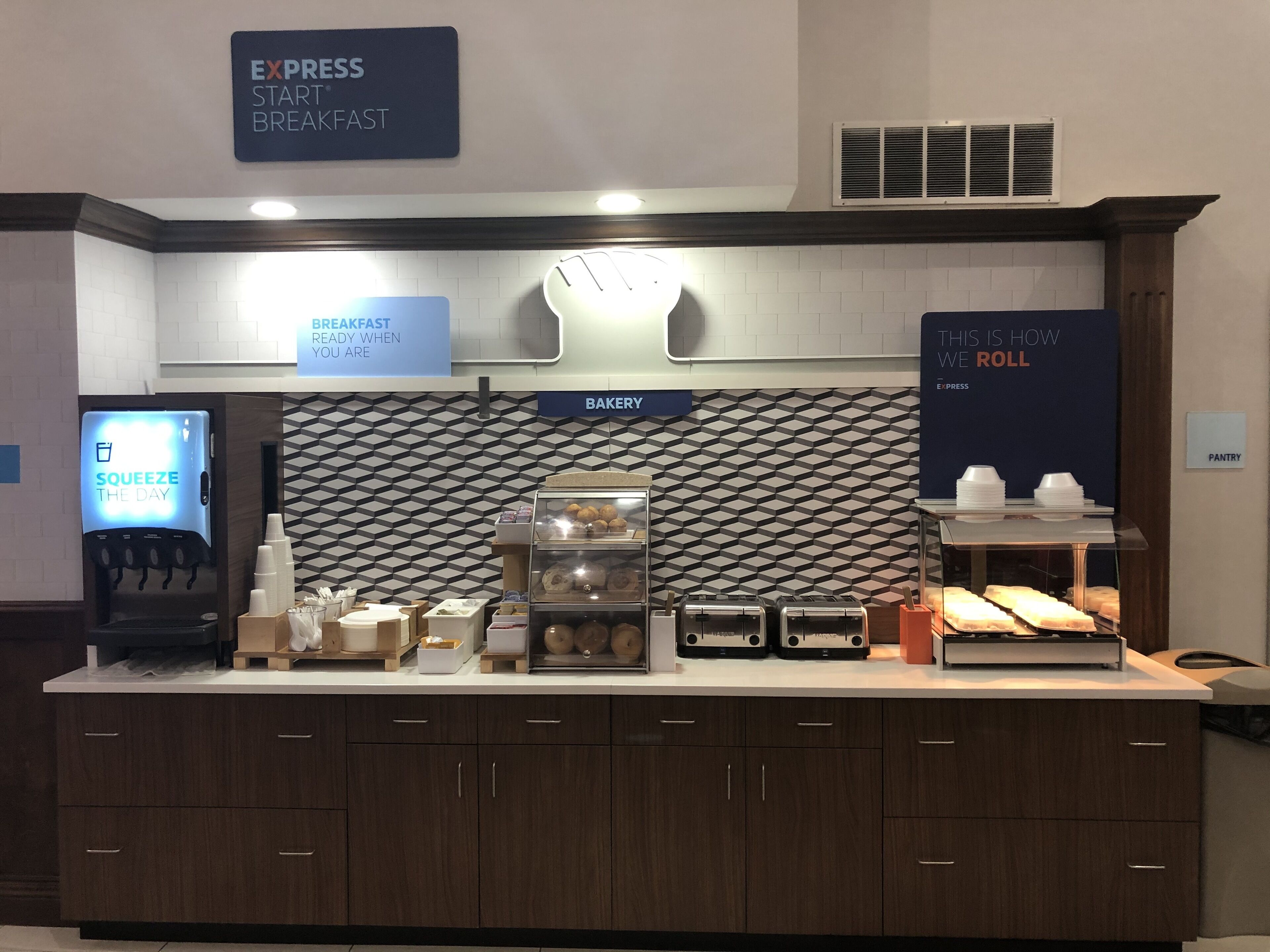 Un espace dédié au petit-déjeuner, équipé d’une machine à café en libre-service, d’un présentoir de produits de boulangerie et d’un panneau indiquant que le petit-déjeuner est servi.