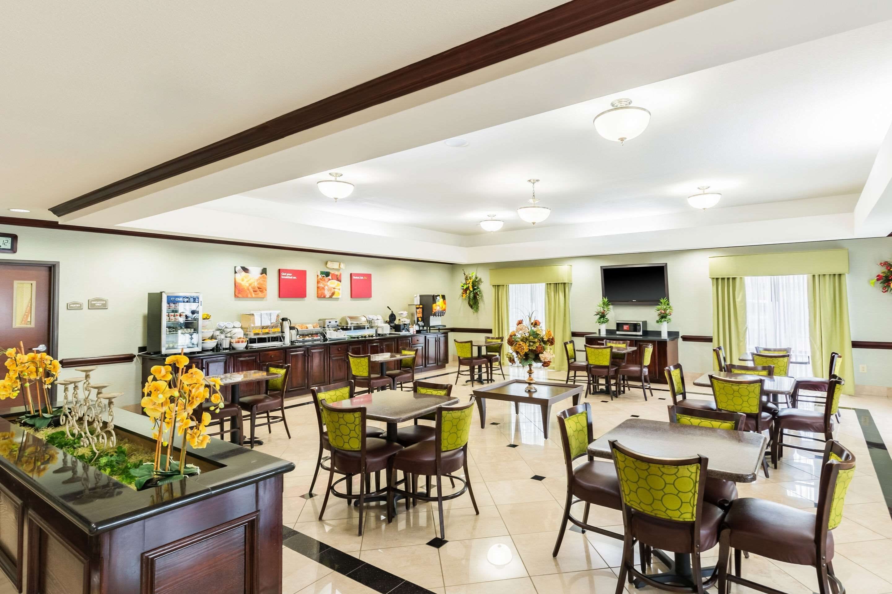 A spacious hotel dining area with multiple tables and chairs, a central counter with a display of food items, and a kitchen area in the background.