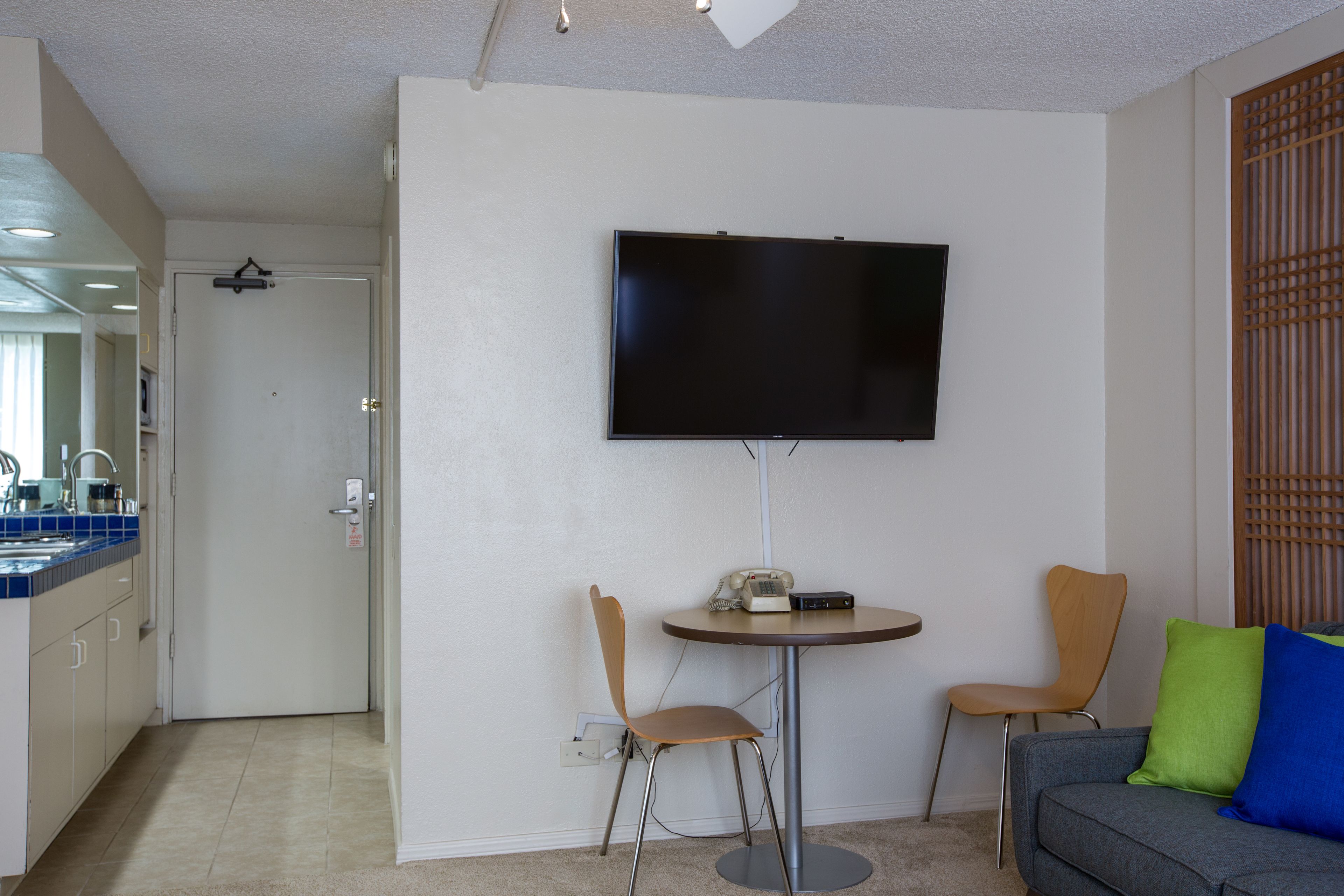Ett modernt vardagsrum med en platt-TV som är monterad på väggen, en grå soffa med blå och gröna kuddar och två trästolar.