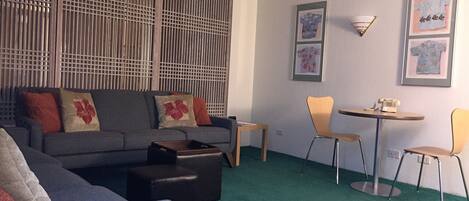 A living room with a grey sofa, a coffee table, and a small side table. There are two chairs, a wall-mounted lamp, and framed artwork on the wall.