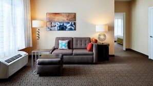A hotel room with a sofa, ottoman, two wall-mounted lamps, a heater, and a patterned carpet. - TownePlace Suites Harrisburg Hershey (Harrisburg)