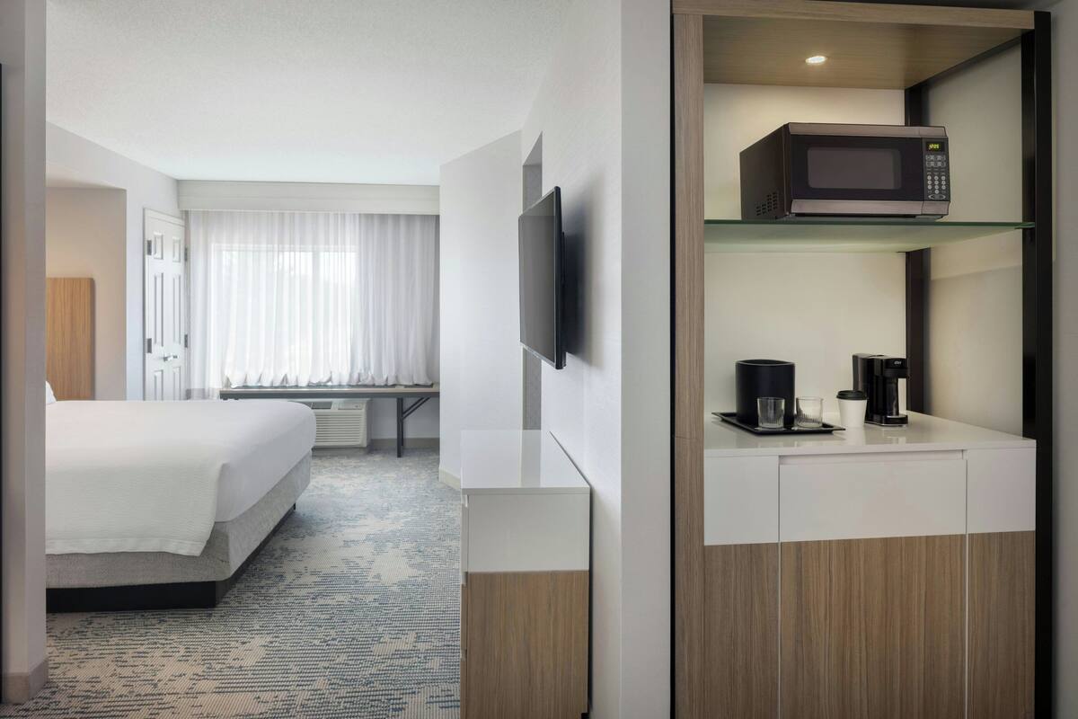 A hotel room with a bed, a flat-screen TV mounted on the wall, a microwave on a shelf, a coffee maker, and a window with curtains.