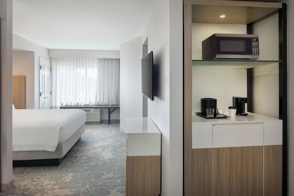 Habitación de hotel con cama, televisor de pantalla plana montado en la pared, microondas en un estante, cafetera y ventana con cortinas.