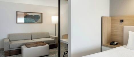 A hotel room with a sofa, ottoman, bedside table, lamp, and a framed abstract painting on the wall.