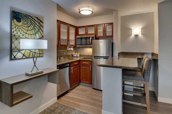 A compact kitchen with wooden cabinets, a stainless steel refrigerator, a microwave, and a built-in island with a chair. - Staybridge Suites Missoula by IHG (Missoula)