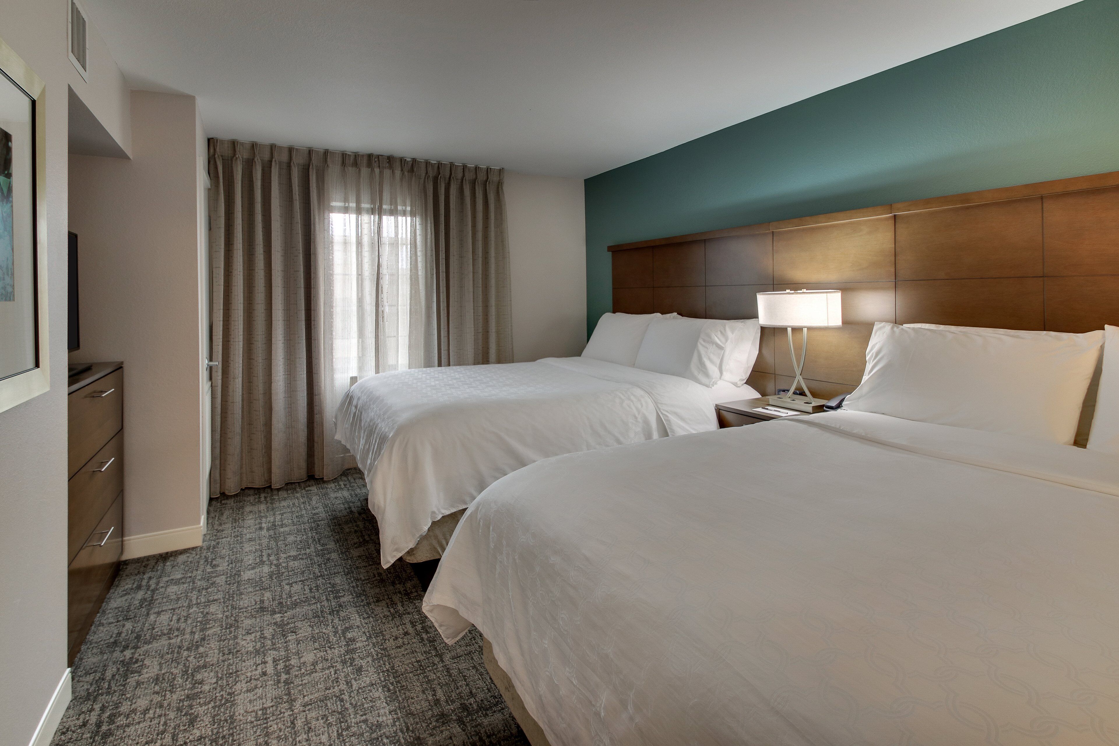 Une chambre d’hôtel avec deux lits, une tête de lit en bois, une table de chevet avec une lampe, une commode et une fenêtre avec des rideaux.