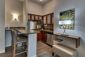 A compact kitchen with wooden cabinets, a stainless steel refrigerator, a microwave, and a dishwasher. There is a bar area with a cushioned chair and a small shelf. - Staybridge Suites Missoula by IHG (Missoula)