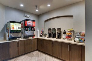 A self-service beverage station with multiple coffee makers, a Coca-Cola dispenser, and a fruit display.