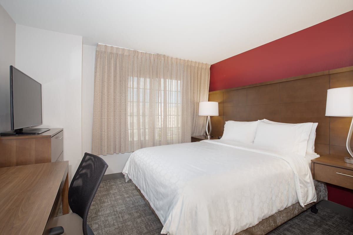 A hotel room with a bed, a desk with a television, a chair, a nightstand with a lamp, and a window with curtains.