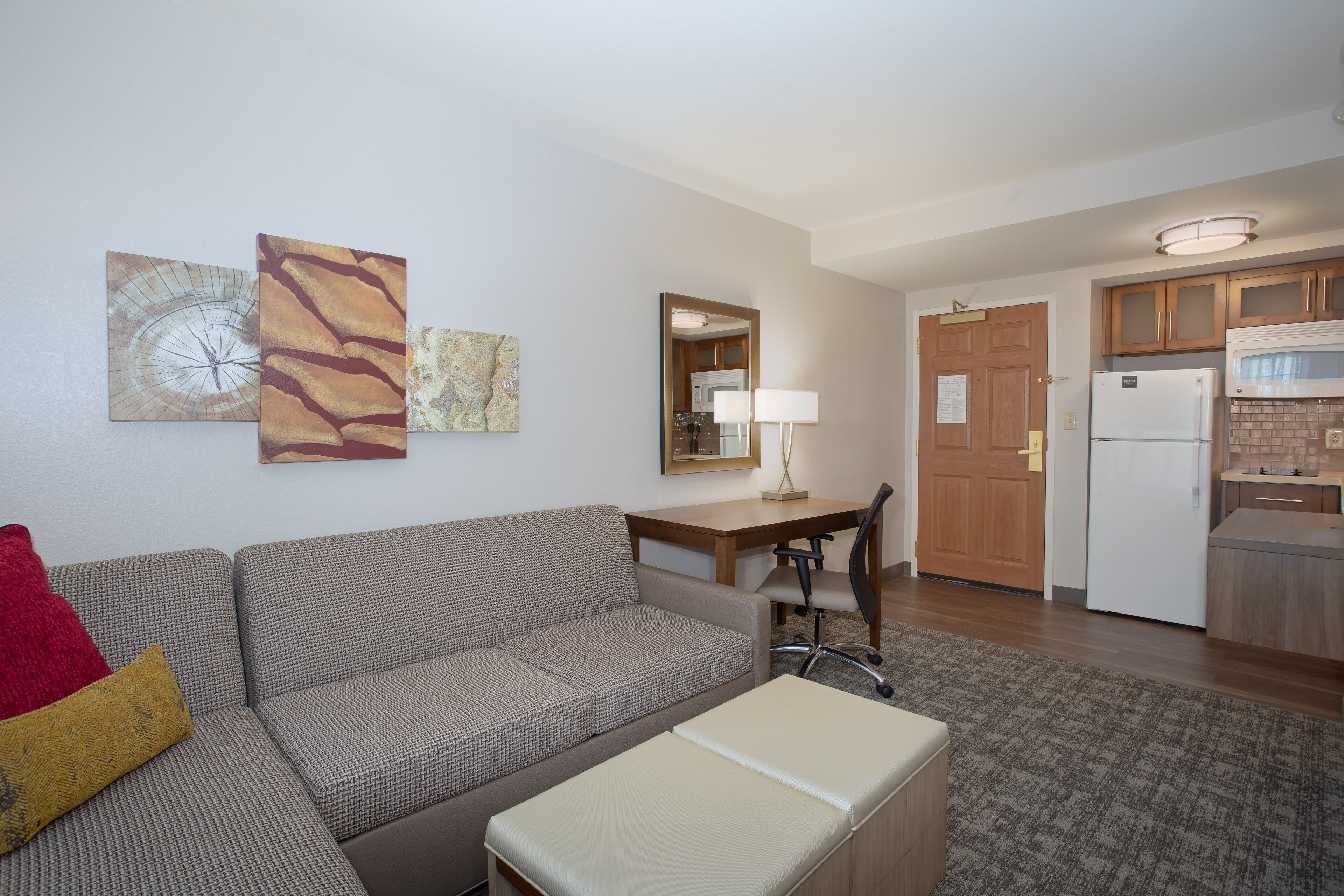 A modern living room with a sofa, a small table, and a lamp. There is a kitchen area with cabinets and a refrigerator.