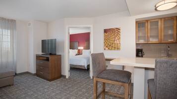 A hotel room with a bed, a desk with a chair, a television, a kitchen area, and a painting on the wall.