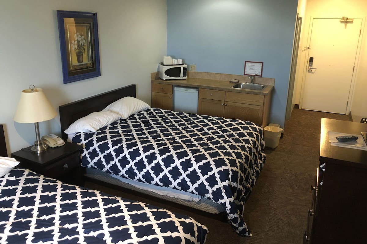 A hotel room with two beds, a bedside table with a lamp, a microwave, a small sink, and a door leading to another room.