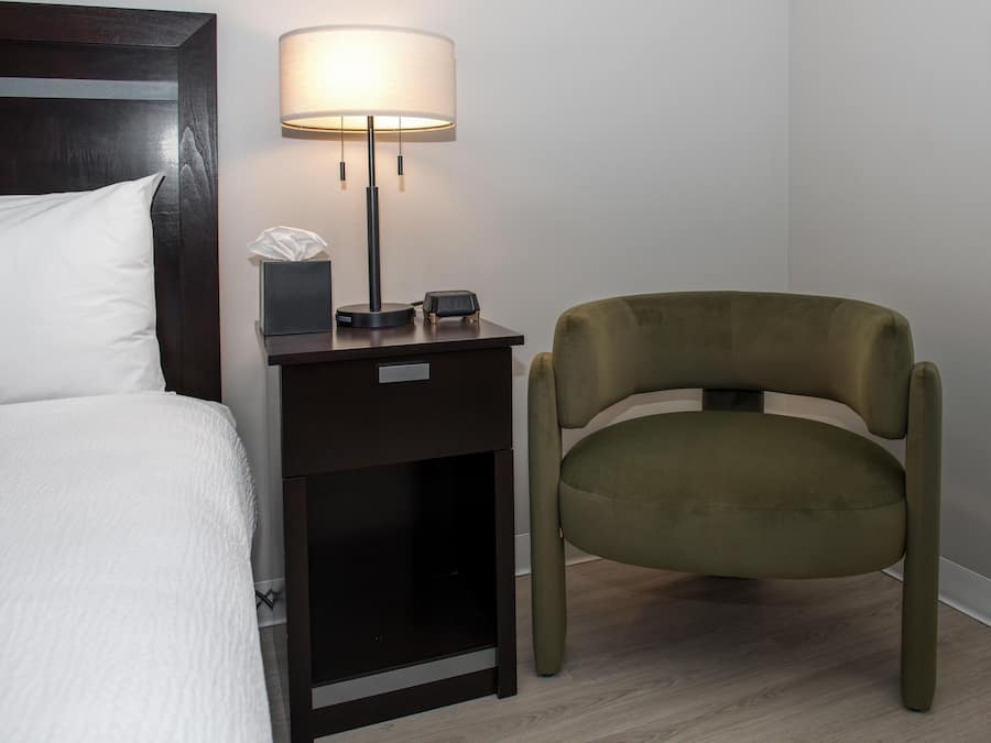 Una cama bien hecha con edredón blanco, una mesita de noche con lámpara, caja de pañuelos y reloj digital.