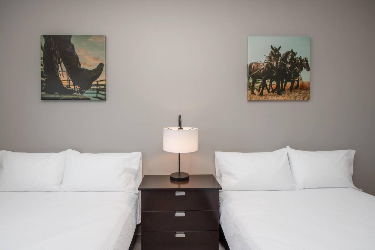 A hotel room with two beds, a nightstand with a lamp, and two paintings on the wall.