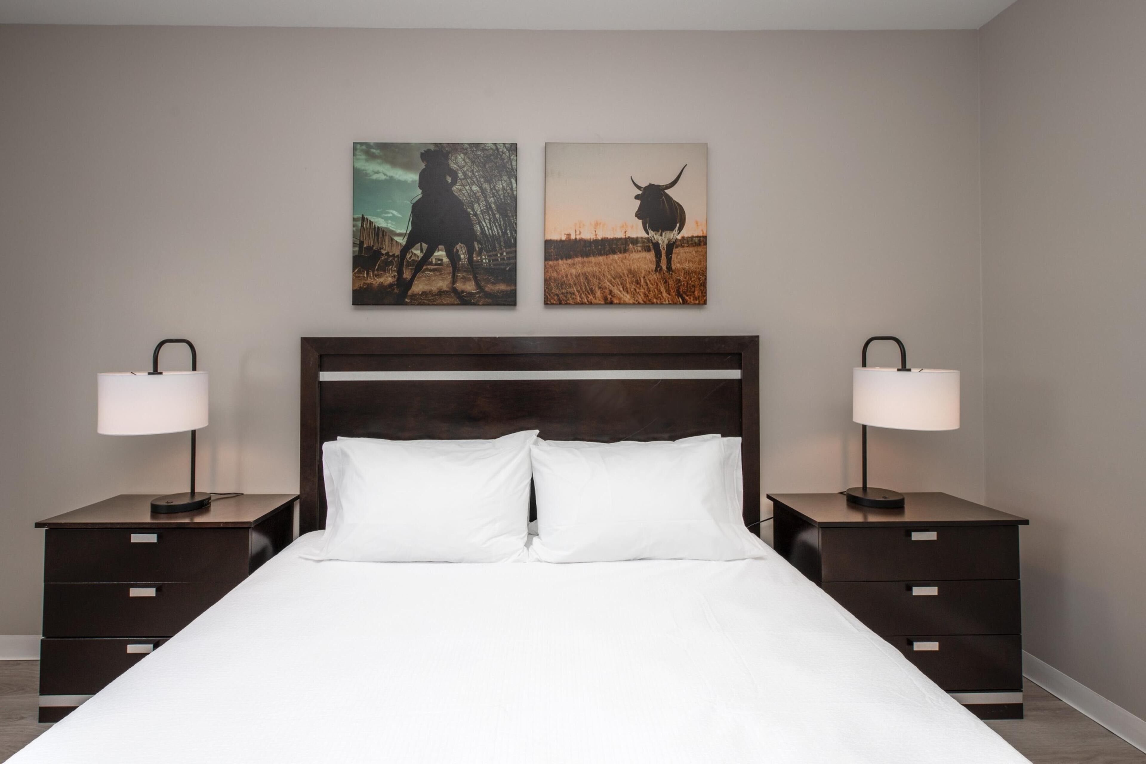 Une chambre d’hôtel avec un grand lit, deux tables de chevet, deux lampes fixées au mur et deux tableaux encadrés.