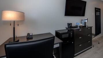 Habitación de hotel con escritorio, silla, lámpara, televisión de pantalla plana, minibar y cafetera.