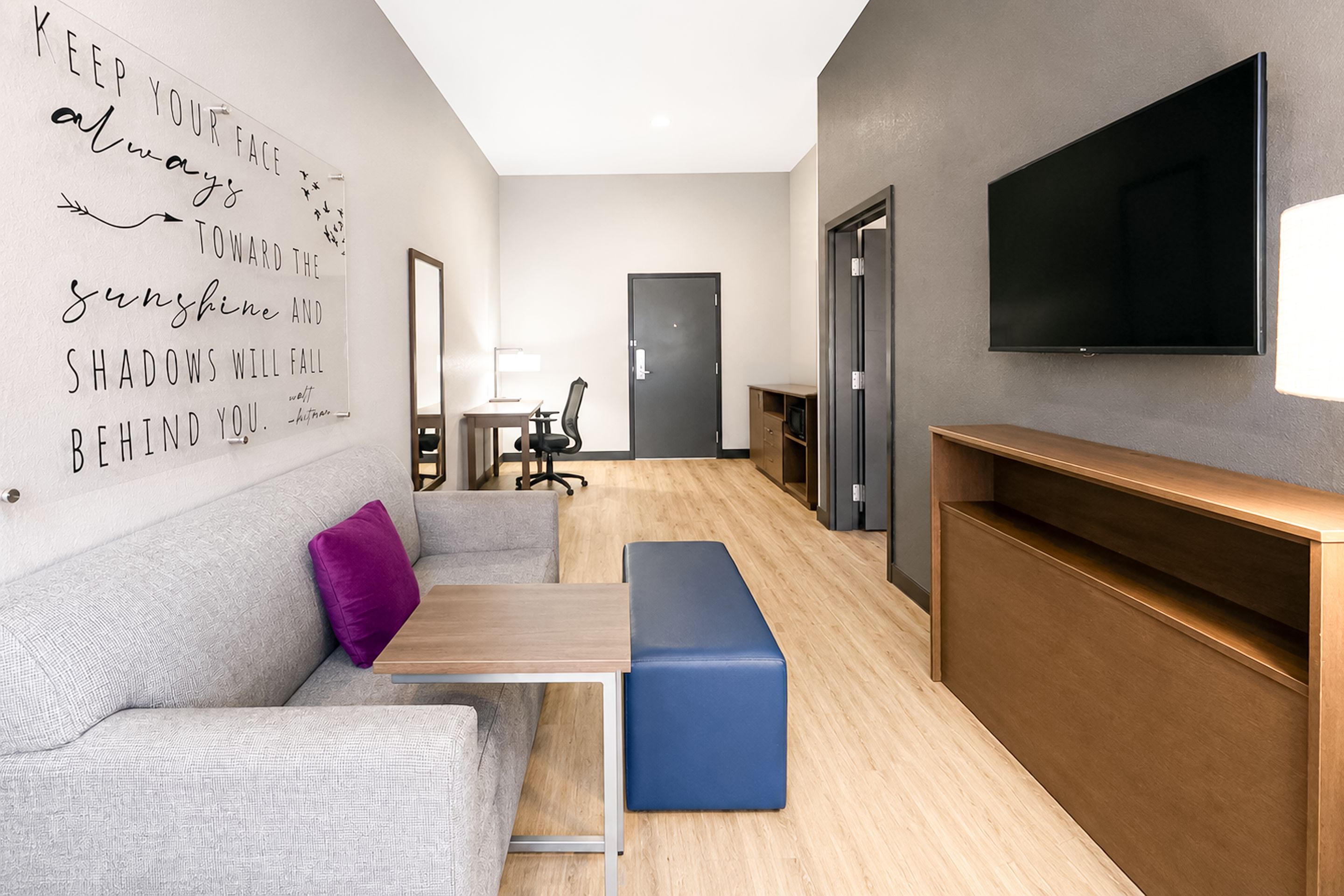 A modern hotel room with a sofa, a desk, a flat-screen TV, and a large mirror.