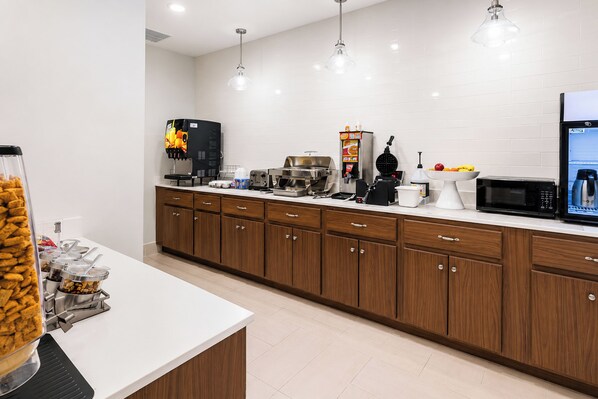A modern kitchen with a white countertop, wooden cabinets, and various appliances like a coffee machine, waffle maker, and microwave. - La Quinta Inn & Suites by Wyndham Augusta/Fort Eisenhower (Augusta)