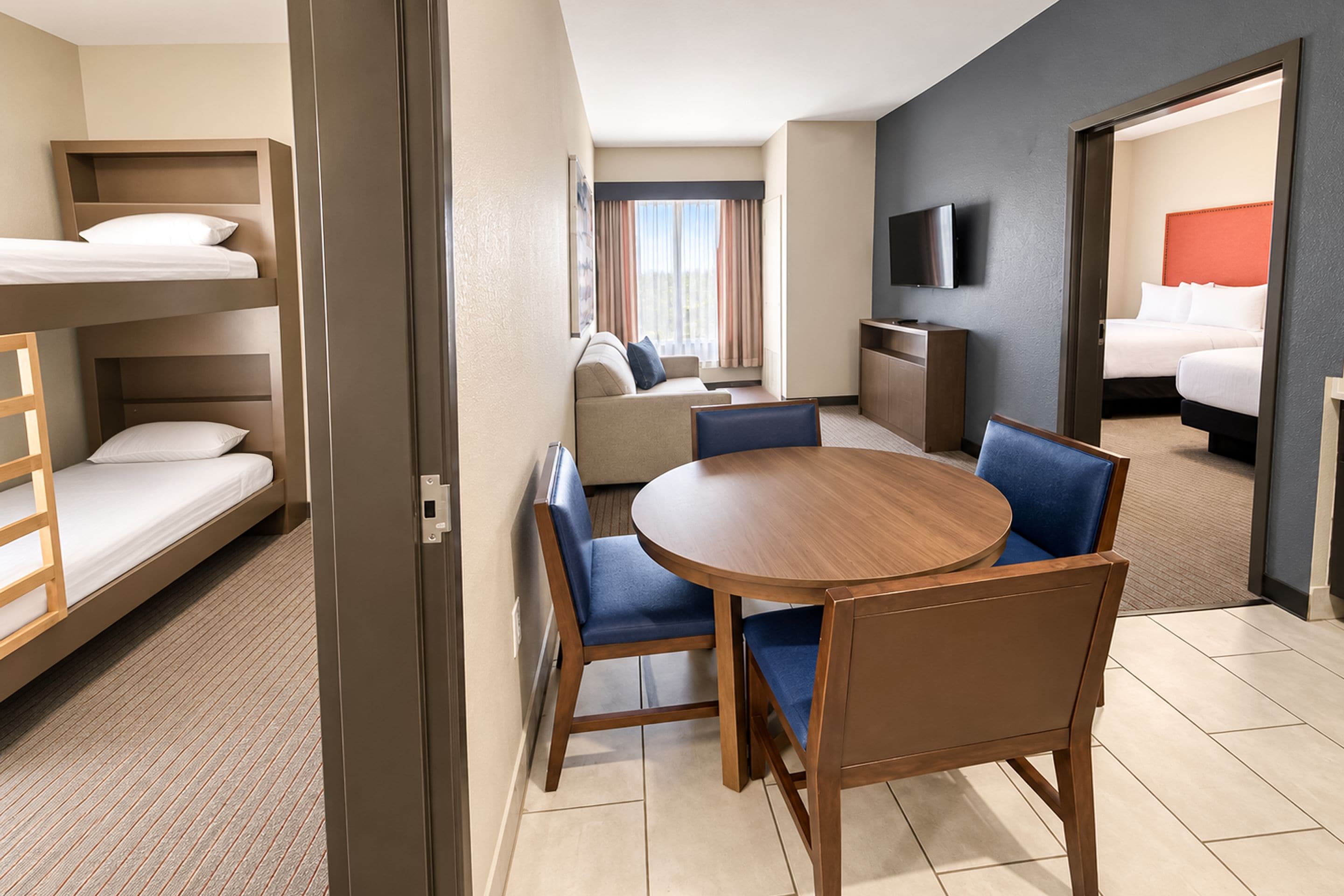 A hotel room with a bunk bed, a round dining table with chairs, a sofa, and a television.