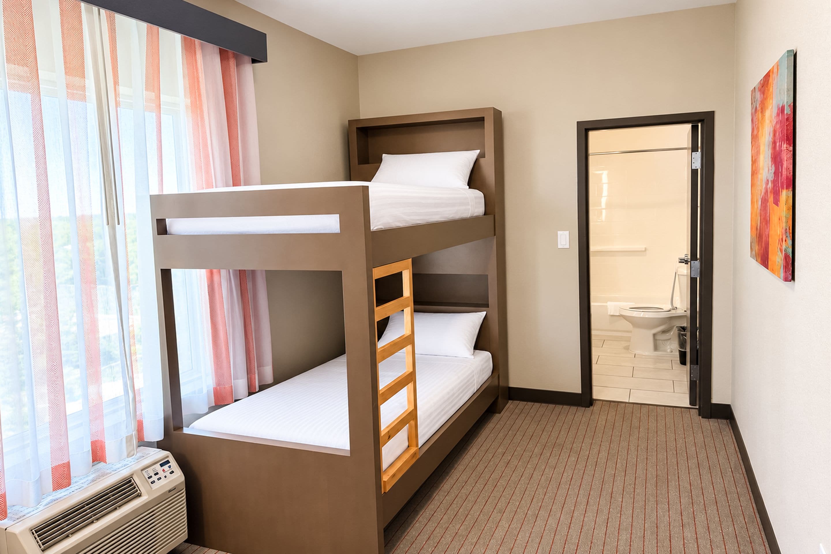 A bunk bed room with a window, curtains, an air conditioning unit, and a bathroom visible through an open door.