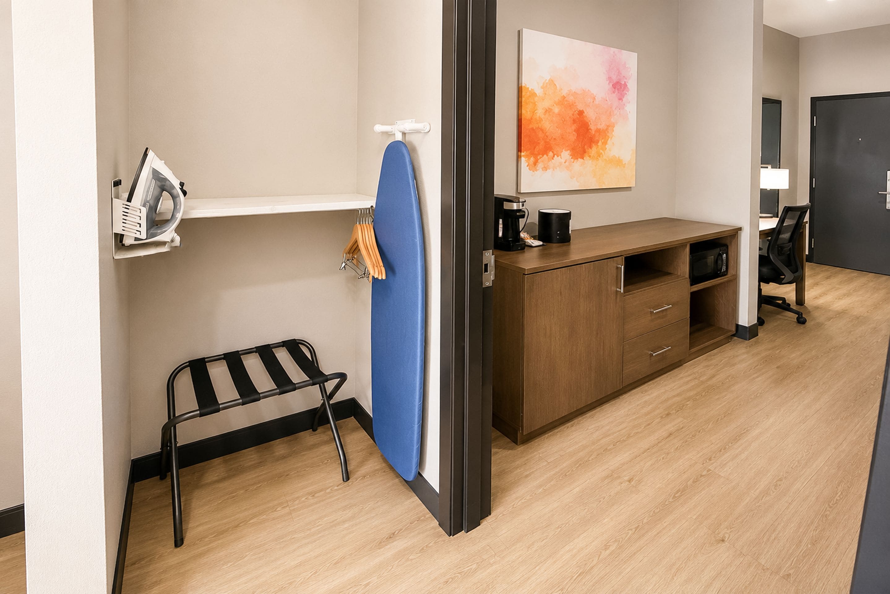 A hotel room with a wooden desk, a coffee maker, a microwave, a chair, and a painting on the wall.