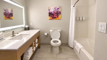 Un baño con diseño moderno, que incluye un lavamanos, inodoro y bañera. Hay toallas y artículos de aseo en el mostrador.