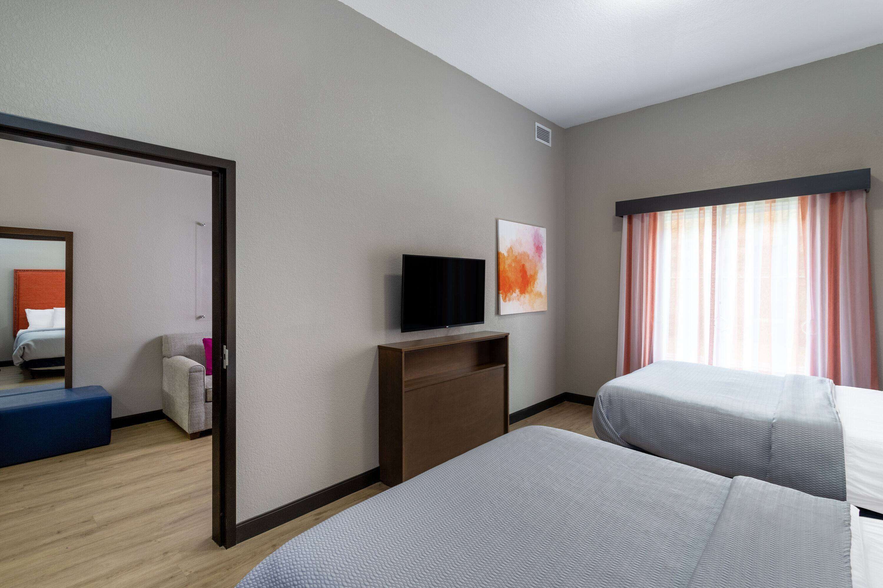 Camera d'albergo con due letti, una TV su un mobile in legno, un quadro appeso al muro e una finestra con tende.
