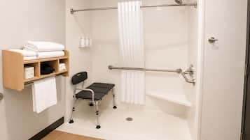 Zona de ducha con barra de apoyo, silla de ruedas y estante con toallas.