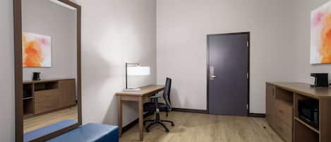 A hotel room with a wooden desk, a chair, a blue bench, a microwave, a coffee maker, and a door.