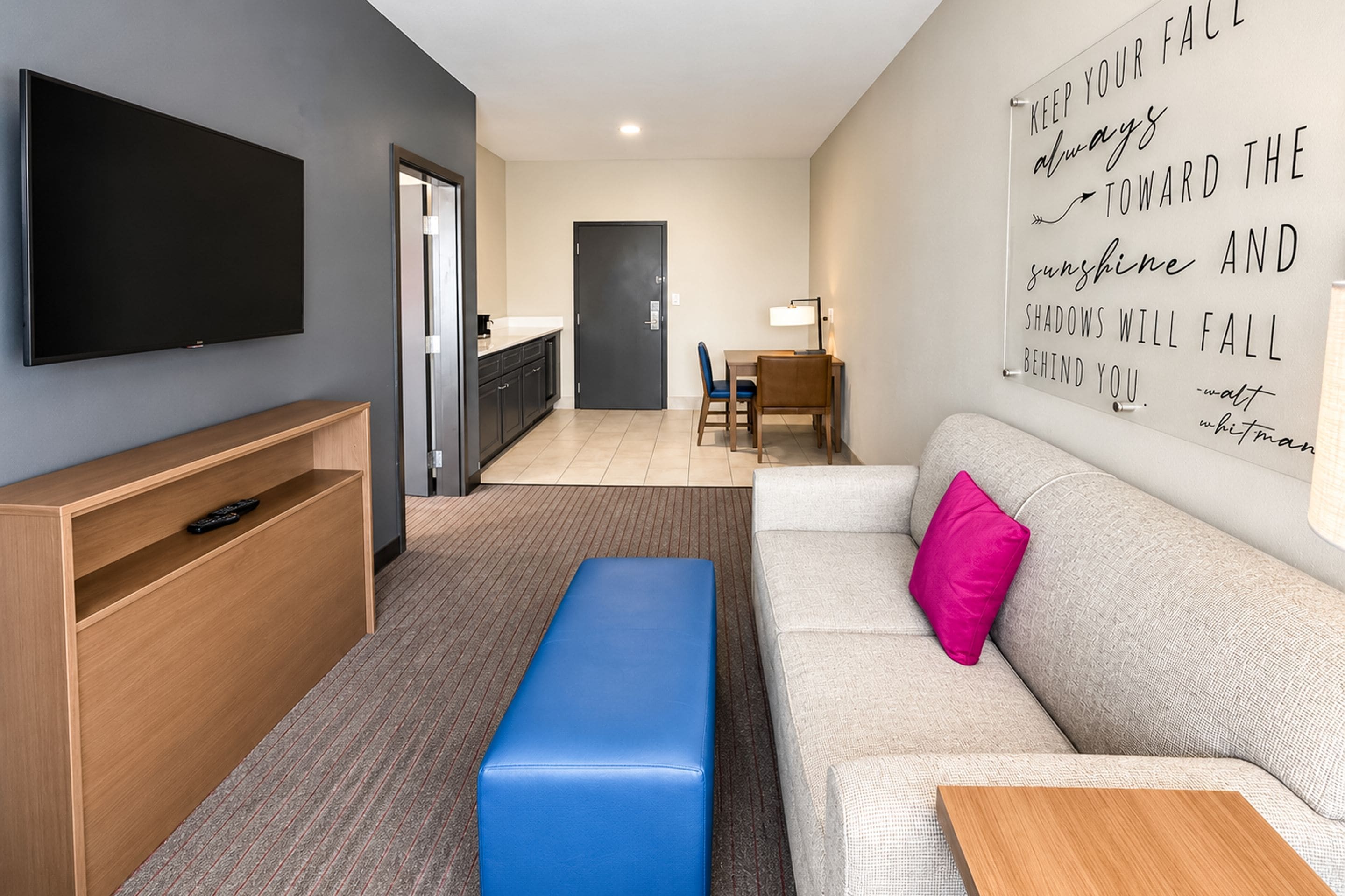 Un salon moderne doté d’un téléviseur à écran plat, d’un banc en bois, d’un canapé agrémenté d’un coussin rose vif et d’une citation inspirante sur le mur.