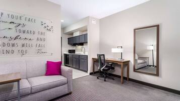 A hotel room with a grey sofa, a desk with a chair, a kitchen area, and a large mirror on the wall.
