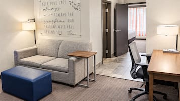 Een hotelkamer met een bank, een klein tafeltje, een blauwe poef, een bureau met een stoel, en een muur met een motiverende quote.