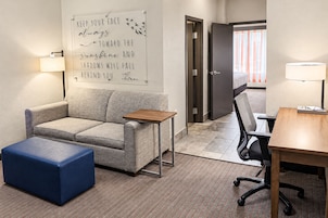A hotel room with a sofa, a small table, a blue ottoman, a desk with a chair, and a wall with a motivational quote.