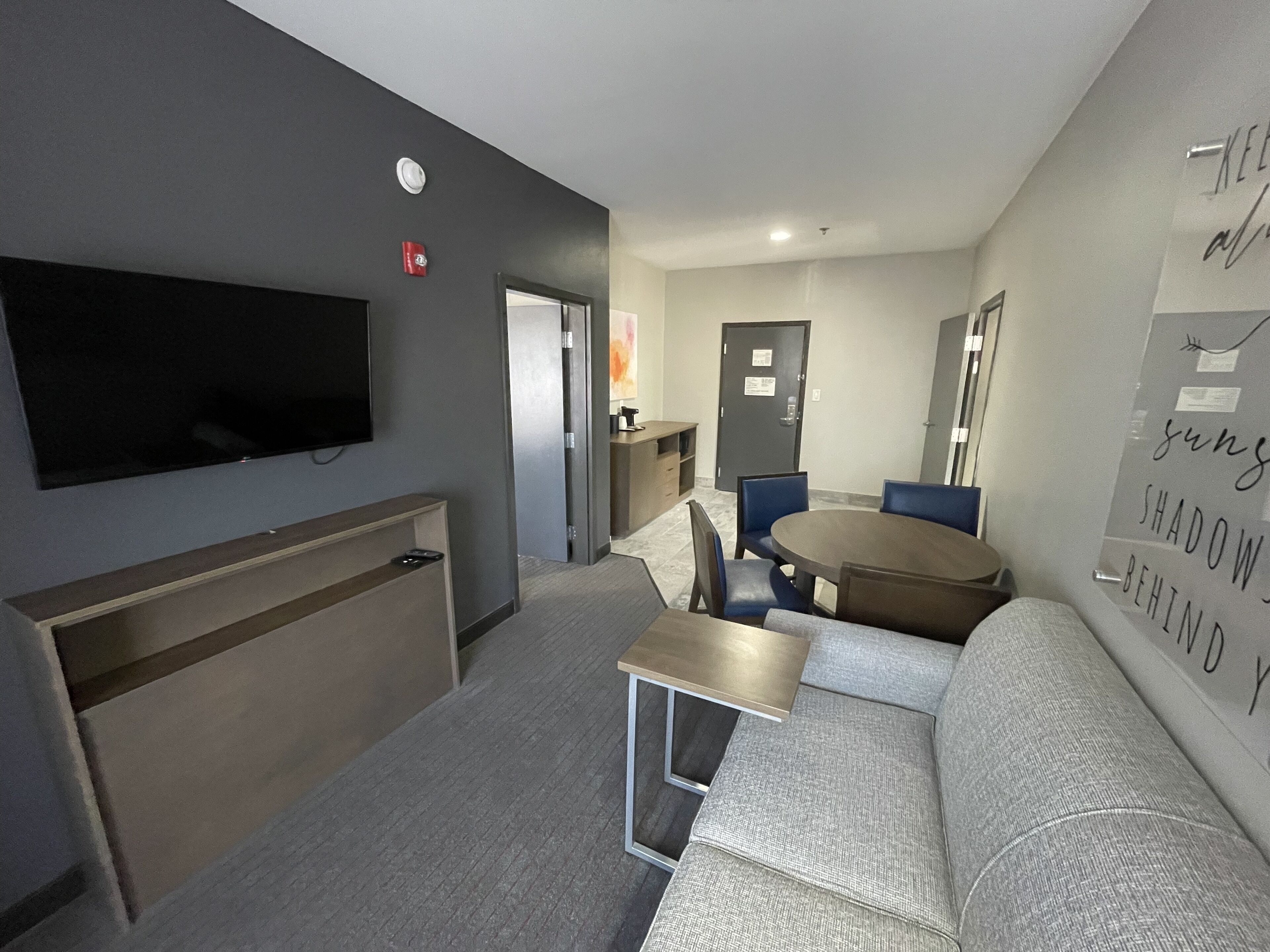 A modern hotel room with a flat-screen TV, a sofa, a dining table, and a small kitchenette.
